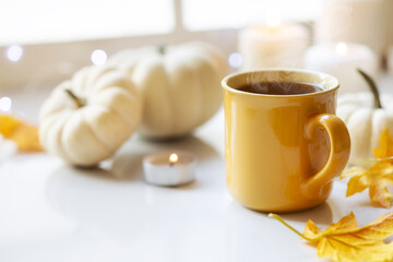 Cozy Autumn setting with steaming cup of tea, white pumpkins and autumnal leaves on a window sill at the background