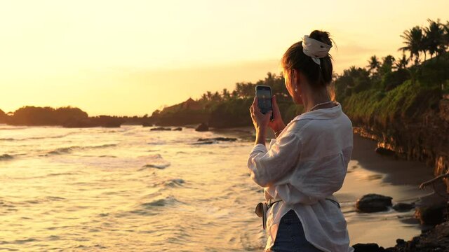 Smiling caucasian woman blogger holding mobile phone taking picture of scenic sunset over ocean during vacation trip, positive hipster girl influencer shooting video on smartphone standing on beach
