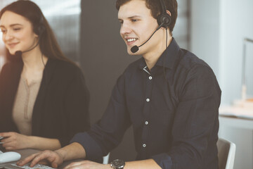 Young dark-haired guy with headsets is talking to a client, while sitting in sunny office. Call center operators at work