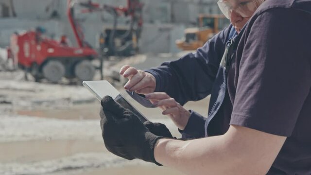 Handheld mid-section shot of female supervisor in safety goggles gesticulating and explaining something to worker with tablet in granite quarry. Heavy machinery in background
