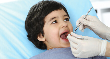Cute arab boy sitting at dental chair with open mouth during oral checking up with doctor. Visiting...