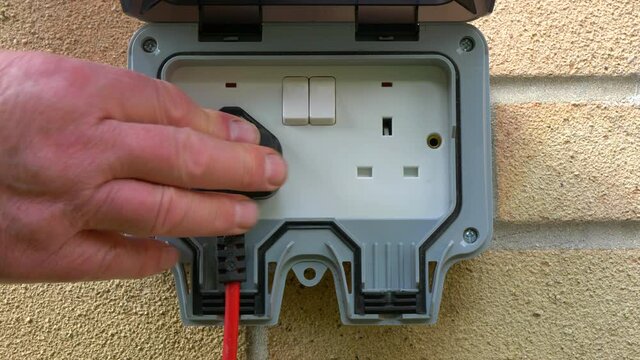 Closeup POV shot of a man’s hand inserting a UK plug on the end of a cable, into a weatherproof, three pin double socket, attached to an outside brick wall, then switching on the electrical supply.