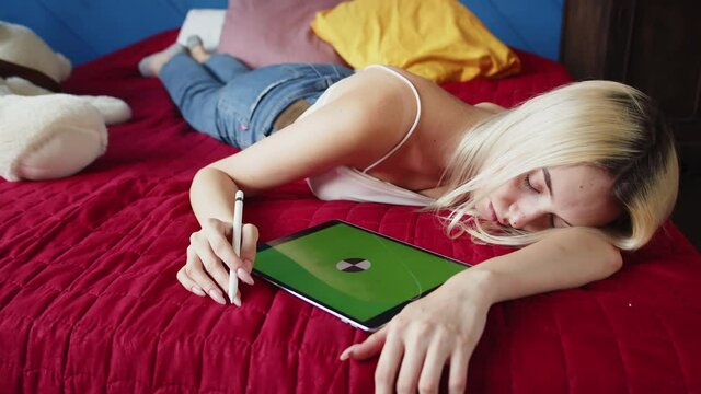 Tired blonde woman is lying on bed and sleeping, holding stylus in her hands and next to it is digital tablet with green screen, mockup, chromakey