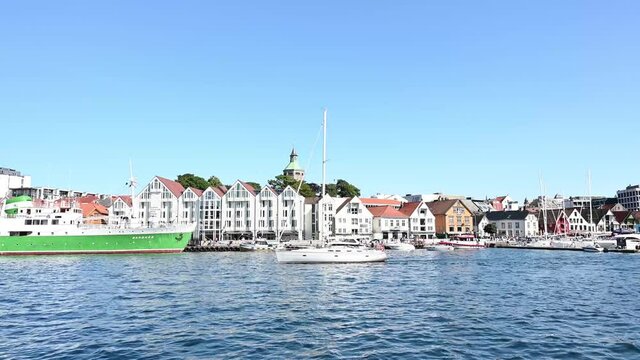 A white sailboat sails through the harbour of Stavanger, Norway on a cloudless summer day.