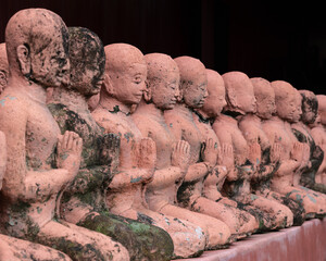 Row of old terracotta statues of monks in prayer on dark background at ancient Wat Phan Tao...