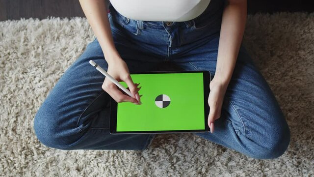 Woman sits in lotus position and draws with stylus on electronic tablet with green screen, top view. Mockup. Concept of digital creativity art