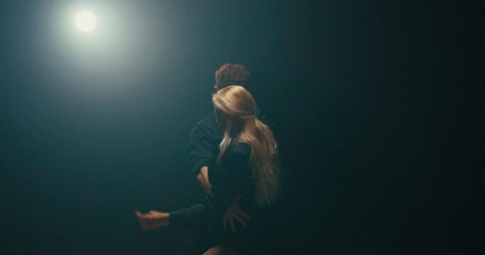 handsome man in an unbuttoned black shirt dances romantically passionately with beautiful blonde with long hair they move their hips woman twists buttocks dark studio scene lit by strong lamp