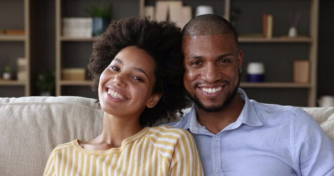 Head shot beautiful smiling African 35s spouses sit on sofa in living room look at camera. Happy marriage, homeowners couple, bank loan, family counselling services clients, harmonic relation concept