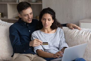 Happy bonding relaxed young family couple holding credit bank card in hands, making payments purchasing goods or services in internet store or shopping app on computer, resting on comfortable sofa.