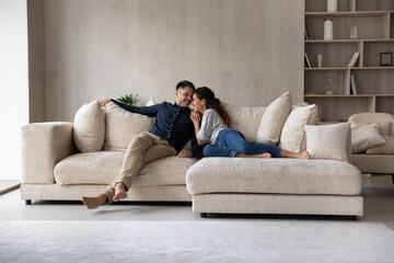 Full length happy relaxed young family couple homeowners or renters lying on big cozy sofa,...