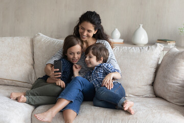 Happy bonding young latin mum and cute kids siblings posing for selfie photo on cellphone or recording streaming live video stories in social network, resting together on sofa, tech addiction.
