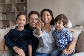 Portrait of smiling young hispanic family couple and cute little children showing keys, sitting on cozy sofa, feeling excited of moving into new own apartment, celebrating purchasing new dwelling.