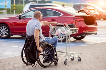 Fototapeta na wymiar Adult disabled man in a wheelchair pushes a cart towards a car in a supermarket parking lot