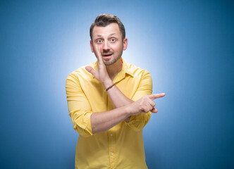 Young guy holding hand near mouth and telling secret over blue background, dresses in yellow shirt. Gossip concept