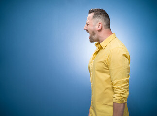 Profile of aggressive young man screaming, copy space for text over blue background, dresses in yellow shirt