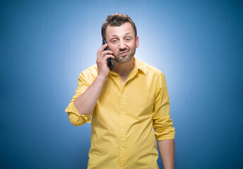 Grumpy man talks on the phone over blue background, dresses in yellow shirt. Nuisance and smartphone