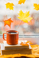 Naklejka na ściany i meble Autumn still life with a beautiful bokeh. Outside the window, maple leaves and raindrops are running down the glass. A cup of hot tea, books, and a knitted blanket by the window.