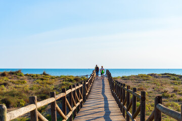 Two unrecognizable people with backpacks walking with a dog along a wooden walkway on the beach, with the sea in the background. 