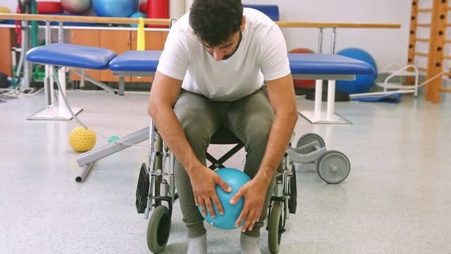 Handheld shot of a Caucasian disabled man seated in a wheelchair, doing an exercise with a smaller pilates ball at the physiotherapeutic rehabilitation center