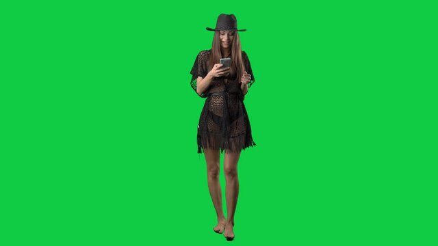 Young slim woman in beachwear with beach tunic dress walking and using cell phone. Full body on green screen chroma key background