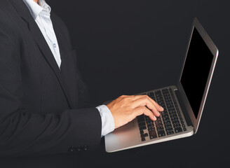 Midsection of a businessman in a suit holding and using a laptop while standing with black background in the studio. Space for text. Business and technology concept