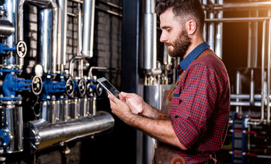 Young male brewer in leather apron supervising the process of beer fermentation at modern brewery factory