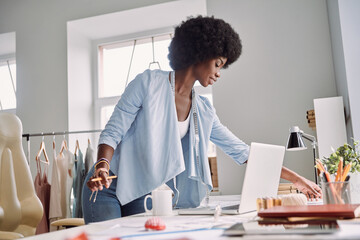 Beautiful young African woman working while standing ner her desk in fashion design studio