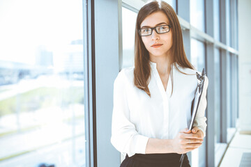 Fototapeta na wymiar Beautiful female specialist with clipboard standing in modern office and smiling charmingly. Working on design, data analysis, plan strategy. Business people concept