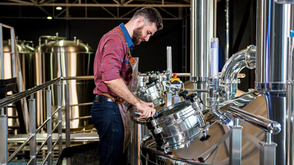 Young male brewer in leather apron supervising the process of beer fermentation at modern brewery...
