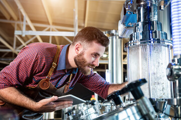 Young male brewer in leather apron supervising the process of beer fermentation at modern brewery factory