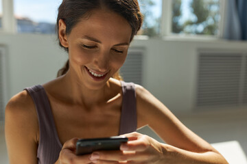 Close-up portrait of sporty slim woman with cute clean face using applications on mobile phone, watching mems, smiling widely showing white healthy teeth, having rest after training