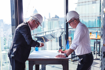 Picture of two engineer  working together in their construction project. They are wearing white shirt , suit and hardhat. They are drawing on a paper with pen in their hand. They are in a hotel lobby.