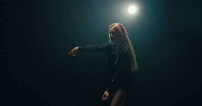 Beautiful experienced talented woman with long blonde hair is dancing on dark stage lit by lamp, practicing new moves, poses, moving sexily, being choreographer, showing steps to beginner dancers