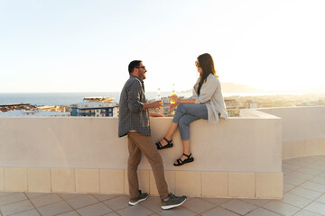 Couple enjoying seascape sunset, drinking beer and eating pizza on the rooftop. Happy people spend quarantine time, picnic leisure on the roof and watch the seaside back view.