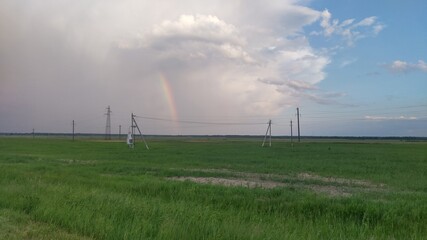 A thundercloud is moving over a sown field with power line poles standing on it. It is raining and a rainbow is visible.