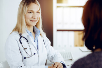 Friendly female doctor and patient woman discussing current health examination while sitting in sunny clinic. Perfect medical service in hospital - 459278067