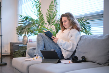 girl sits on the couch and reads a book, a tablet and headphones lie next to it. modern teenager at home. teen literature concept.