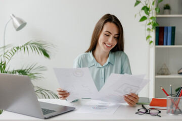 Smiling fun young successful employee business woman in blue shirt hold paper account documents sit work at workplace white desk with laptop pc computer at office indoors. Achievement career concept.
