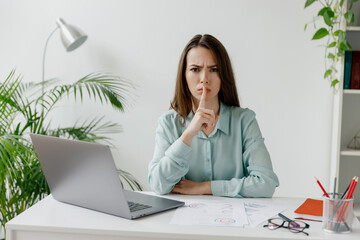 Young employee business woman in blue shirt say hush be quiet with finger on lips shhh gesture sit...