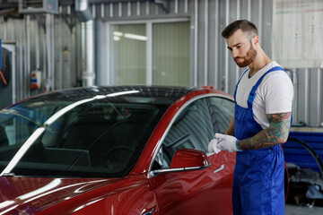 Serious minded young male professional technician car mechanic man 20s wear denim blue overalls white t-shirt gloves stand near red automobile work in light modern vehicle repair shop workshop indoor