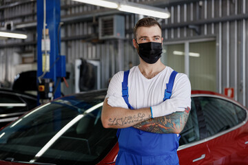 Young professional car mechanic man 20s wears denim blue overalls white t-shirt gloves mask stand...