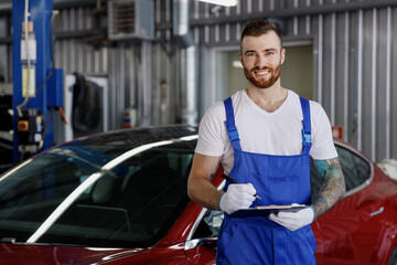 Young happy male professional car mechanic man 20s wears denim blue overalls white t-shirt gloves write hold clipboard with papers document work in light modern vehicle repair shop workshop indoors