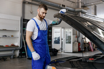 Strong serious confident young male professional technician car mechanic man 20s wears denim blue overalls white t-shirt raised hood bonnet work in light modern vehicle repair shop workshop indoors.