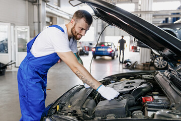 Young fun professional technician car mechanic man in denim blue overalls white t-shirt gloves fixing problem with raised hood work in modern vehicle repair shop workshop indoor Tattoo translate fun.