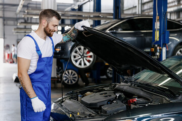 Fun troubleshooter young male professional technician car mechanic man in denim blue overalls white t-shirt fixing problem with raised hood bonnet work in modern vehicle repair shop workshop indoor.