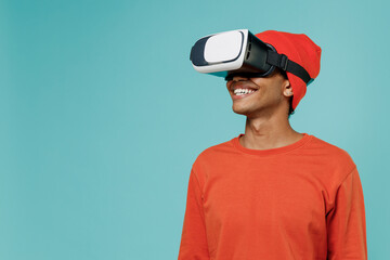 Young cheerful smiling happy african american man 20s in orange shirt hat watching in vr headset pc gadget isolated on plain pastel light blue background studio portrait. People lifestyle concept