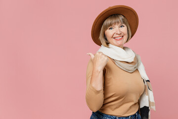 Traveler tourist mature elderly senior woman 55 years old wears brown shirt hat scarf point back on workspace area copy space mock up isolated on plain pastel light pink background studio portrait