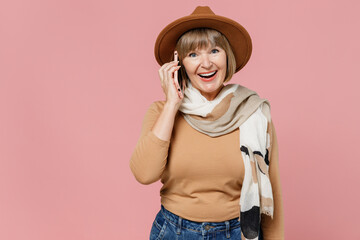Traveler tourist excited fun mature elderly senior lady woman 55 years old wears brown shirt hat scarf talk speak on mobile cell phone isolated on plain pastel light pink background studio portrait.