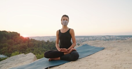 Fototapeta na wymiar Young caucasian woman wearing protective mask sitting in yoga posture and meditating at the mountains. Girl performing morning meditation at the nature. Physical practice concept