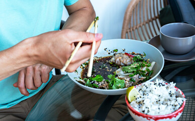 Man in blue t-shirt eating chopsticks healthy vegan food jiaozi with tofu and vegetables in soy sauce with parsley and cilantro and rice with sesame in cafe, chinese dish